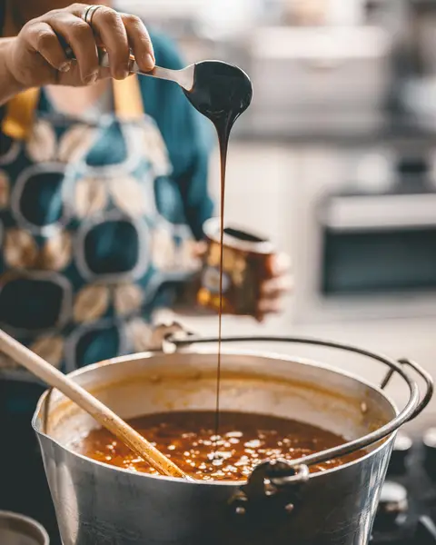 A cook lifts a spoonful of thick homemade jam over a large pot on the stove.