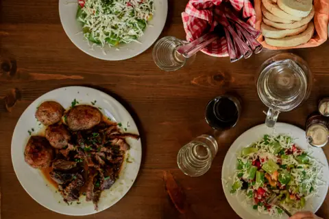 A wooden table seen from above with a plate of slow-cooked meat and roast potatoes, a salad topped with grated cheese, a basket of toasted bread and glasses of water.
